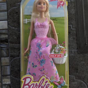 May include: A Barbie doll dressed as an Easter princess in a pink dress with butterflies and flowers. She is holding a basket with a bunny and Easter eggs. The packaging says "Only at Target" and "Barbie Easter Princess".
