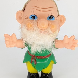 Puede incluir: Un muñeco de gnomo de plástico vintage con un atuendo verde y amarillo, barba blanca y ojos azules. El gnomo lleva un cinturón marrón y zapatos negros.