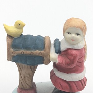 Puede incluir: Figura de cerámica de una niña con abrigo rojo y ribete blanco, arrodillada junto a un buzón azul. Un pájaro amarillo está encima. La base está pintada para parecer nieve. La figura es una decoración navideña.