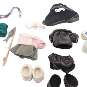 Puede incluir: Colección de ropa y accesorios para muñecas, que incluye un chaleco verde, pantalones cortos, una caña de pescar, un vestido rosa y gris, una chaqueta de cuero marrón, un vestido blanco con una estrella de David azul y zapatos. Fondo blanco.