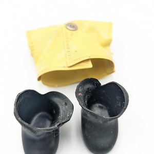 Puede incluir: Un par de botas de goma negras con un puño de cuero amarillo, aseguradas con un broche metálico. Las botas tienen un borde superior único en forma de corazón. El puño está doblado.