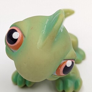 May include: A green toy lizard with large black and orange eyes. The lizard has a rounded head, small legs, and a tail. The toy is made of plastic and has a glossy finish. The lizard is a collectible toy.