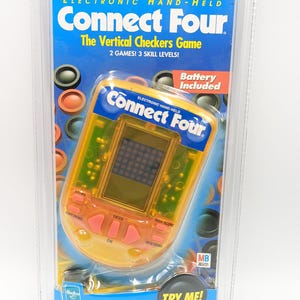 May include: Electronic Connect Four handheld game in a clear plastic package. The game features a yellow and green design with a digital display and buttons. The packaging includes the text "Connect Four" and "The Vertical Checkers Game."
