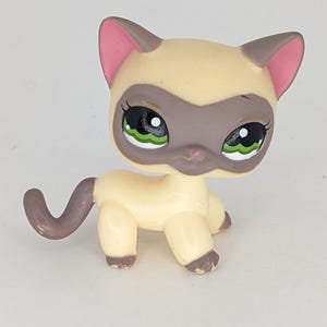 Könnte beinhalten: Eine hellbraune und braune Siamkatzenfigur mit großen grünen Augen. Die Katze hat einen hellbraunen Körper, braune Markierungen auf Gesicht, Ohren und Pfoten sowie einen gebogenen Schwanz. Die Figur ist ein Sammlerspielzeug.