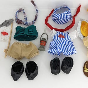 Puede incluir: Un conjunto de diez prendas de ropa para una muñeca pequeña. La ropa incluye un atuendo de salvavidas, un atuendo de pescador, un atuendo de marinero, un atuendo de chef y un atuendo de trabajador de la construcción. La ropa está hecha de tela y tiene varios colores y patrones.