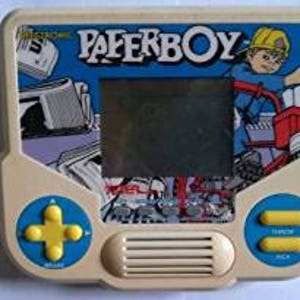 May include: A white handheld electronic game with a blue screen and the words "Electronic" and "Paperboy" printed in black. The game has a yellow directional pad, two yellow buttons, and a speaker.