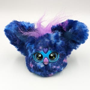 Op de afbeelding: Een pluche speelgoed met een blauw en paars gespikkeld ontwerp. Het speelgoed heeft grote, expressieve ogen, een blauw voorpaneel en paarse plukjes haar. De oren van het speelgoed zijn groot en pluizig, en het heeft een hartvormig detail boven de ogen.