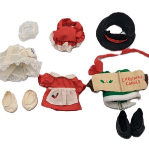 Puede incluir: Un conjunto de ropa y accesorios para muñecas, que incluye un vestido blanco con ribete de encaje, vestidos rojos y blancos, un sombrero negro con una banda roja y un libro con "Christmas Carols". También se incluyen zapatos blancos y negros.