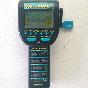 K&ouml;nnte beinhalten: Ein schwarzes und blaues Handheld-Videospiel mit einem Bildschirm, der eine Karte eines Sees und den Text "Bass Fishin'" oben anzeigt. Das Spiel hat Kn&ouml;pfe f&uuml;r "See", "Boot", "Kescher", "Spiel", "Bester Fang", "Sound", "K&ouml;der - Schleppen", "Start - Auswerfen", und "Zur&uuml;cksetzen". Das Spiel hat auch eine blaue Kurbel auf der rechten Seite. Die Unterseite des Spiels zeigt eine Liste von "Tackle Box"-Artikeln, darunter "Kunstk&ouml;der", "Jigs", "Spinnerbaits", "Crankbaits", und "Buzzbaits".