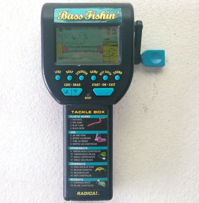 K&ouml;nnte beinhalten: Ein schwarzes und blaues Handheld-Videospiel mit einem Bildschirm, der eine Karte eines Sees und den Text "Bass Fishin'" oben anzeigt. Das Spiel hat Kn&ouml;pfe f&uuml;r "See", "Boot", "Kescher", "Spiel", "Bester Fang", "Sound", "K&ouml;der - Schleppen", "Start - Auswerfen", und "Zur&uuml;cksetzen". Das Spiel hat auch eine blaue Kurbel auf der rechten Seite. Die Unterseite des Spiels zeigt eine Liste von "Tackle Box"-Artikeln, darunter "Kunstk&ouml;der", "Jigs", "Spinnerbaits", "Crankbaits", und "Buzzbaits".