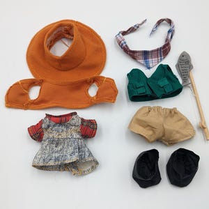 Puede incluir: Un conjunto de ropa de muñeca, que incluye una visera naranja, una bandana a cuadros, un chaleco verde, pantalones cortos color beige, un vestido de mezclilla con mangas a cuadros rojas, zapatos negros y una caña de pescar con un señuelo de pez.