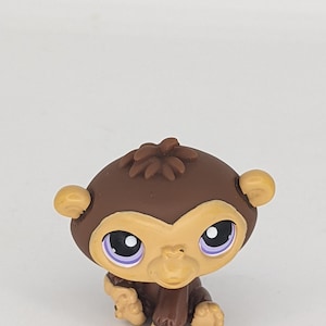 May include: A brown and tan monkey figurine with large, expressive eyes and small ears. The toy monkey is in a sitting position, with its hands and feet visible. The figurine has a detailed, textured design.