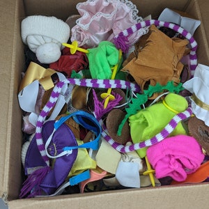 Puede incluir: Una caja de cartón llena de ropa y accesorios para muñecas, incluyendo un sombrero blanco, un vestido rosa, un chaleco marrón, una camisa verde, una falda morada y una blusa blanca.