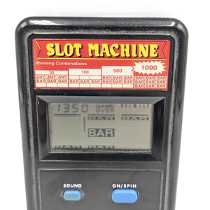 May include: A black handheld electronic slot machine game with a digital LCD screen displaying the text GAME OVER and BAR symbols. The device features a red panel with the text SLOT MACHINE and a chart of winning combinations, plus two blue buttons.