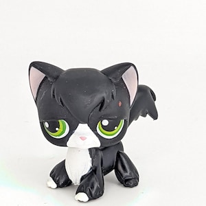 Könnte beinhalten: Schwarz-weiße Katzenfigur mit grünen Augen und rosa Innenohren. Die Katze hat eine weiße Brust und Pfoten sowie einen kleinen roten Fleck im Gesicht. Die Figur ist in einer sitzenden Position mit kleinen Flügeln dargestellt.
