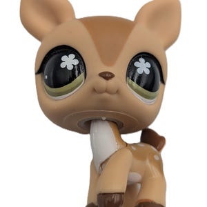 May include: A tan deer figurine with a large head and oversized black eyes featuring white flower-shaped pupils. The deer has a white chest patch, dark brown hooves, and small white spots on its back. It has pointed ears and a small brown nose.