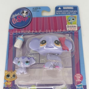 Op de afbeelding: Littlest Pet Shop speelgoedset met een paarse olifant, een baby-olifant en een fles. De olifant heeft een roze strik. De verpakking heeft de tekst "Bobble in Style!" en "Ages 4+".