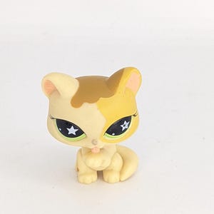 May include: A small, cream-colored cat figurine with brown and yellow markings. The cat has green eyes with white stars and is licking its paw.