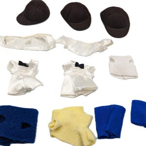 Puede incluir: Un surtido de ropa y accesorios para muñecas, que incluye tres gorras marrones, dos vestidos de satén blanco con lazos negros, un chaleco blanco, pantalones cortos azules y un suéter amarillo. Los artículos están dispuestos sobre un fondo blanco.