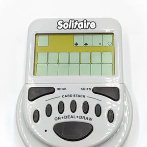 May include: A handheld electronic Solitaire game in a light gray casing. The screen displays a digital card layout, with buttons for deck, suits, card stack, deal, and draw. Additional buttons include game select, new game, high score, and sound.
