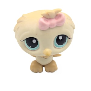 May include: A small, tan-colored bird figurine with a large head and oversized eyes. The bird features a light blue iris, black pupils, and a small, pointed beak. A soft pink bow is attached to the top of its head, and it has two small feet.