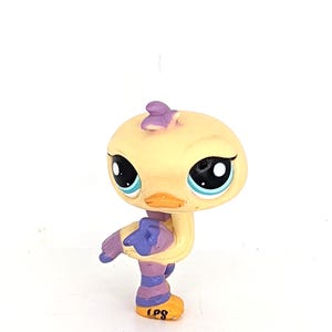 May include: A yellow and purple Littlest Pet Shop duck figurine. The duck has large blue eyes, an orange beak, and purple accents on its head, wings, and legs. The letters "LPS" are printed on the bottom of its feet.