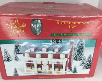 May include: A red box with the words "A Wonderful Holiday" and "Knickerbocker Inn" printed on it. The box contains a miniature illuminated porcelain building of a house with snow-covered roof and trees. The box is a tribute to the movie "It's a Wonderful Life."