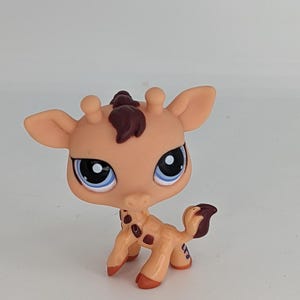 May include: A small, tan giraffe figurine with dark brown spots and a tuft of hair. The giraffe has large blue eyes with white highlights and small ears. The figurine is a collectible toy.