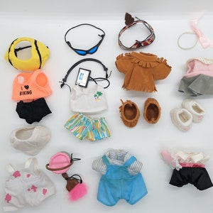 Puede incluir: Una colección de ropa y accesorios para muñecas, que incluye un disfraz de abeja amarillo, un vestido rosa y gris, una chaqueta de gamuza marrón, un conjunto de mono azul, una camisa blanca con un diseño de palmera, un par de zapatos blancos, un par de mocasines marrones, una diadema rosa, un par de gafas de sol azules, un conjunto de vaquera rosa y blanca y una falda floral rosa y blanca.