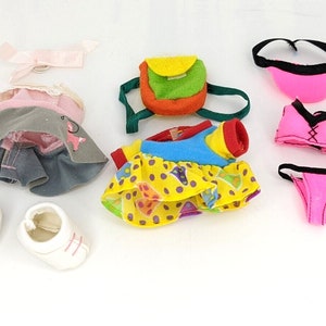 Puede incluir: Un conjunto de ropa para muñecas que incluye un vestido rosa y gris, un vestido amarillo con lunares, un traje de baño rosa, una diadema rosa, una mochila verde y naranja y un par de zapatos blancos.