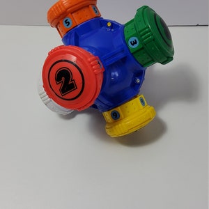 Może przedstawiać: Kolorowa plastikowa zabawka z niebieskim korpusem centralnym i czterema obracającymi się cylindrami w kolorze pomarańczowym, zielonym, czerwonym i żółtym. Każdy cylinder ma numer od 2 do 9. Zabawka przeznaczona jest do zabawy i nauki dzieci.
