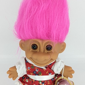 Może przedstawiać: Lalka troll z różowymi włosami, ubrana w czerwoną i białą sukienkę w kwiaty z białą koronkową lamówką. Lalka trzyma koszyk z białym szczeniakiem w środku.