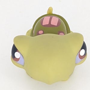 May include: A green and pink plastic toy turtle with large eyes. The turtle has a shell with pink spots and a brown design.