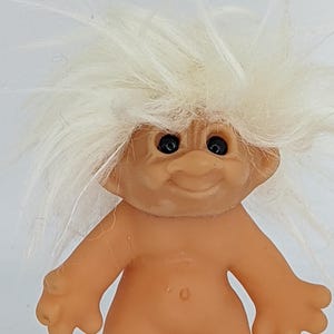 Puede incluir: Una muñeca troll vintage con un cuerpo bronceado, los brazos extendidos y una cara sonriente. La muñeca tiene ojos negros y una gran cabeza de pelo blanco y puntiagudo. Un juguete clásico.