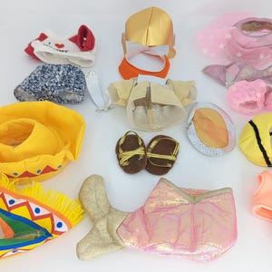 Puede incluir: Una colección de disfraces de tela coloridos y estampados para un animal de peluche. Los disfraces incluyen un sombrero amarillo y rojo, una cola de sirena rosa y dorada, un vestido azul y blanco, un disfraz de abeja amarillo y negro, un disfraz de hada rosa y blanco, un gorro de Papá Noel rojo y blanco, una corona dorada y un traje de baño rosa y blanco.