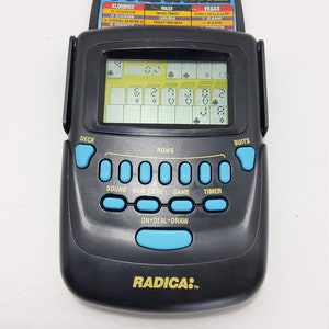 May include: A black handheld electronic game with a blue LCD screen displaying a game of solitaire. The game has buttons for sound, new game, game, timer, and on deal draw. The game is made by Radica.