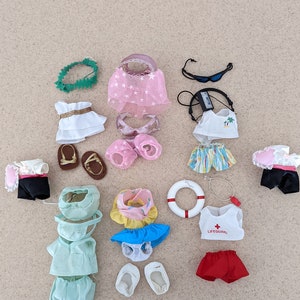 Puede incluir: Una colección de ropa y accesorios para muñecas con temática de playa. Los artículos incluyen un collar de flores verde, un vestido rosa con estrellas, un vestido blanco con cinturón dorado, un top de bikini rosa, una falda a rayas, una falda azul y blanca, una camiseta de salvavidas con una cruz roja, pantalones cortos rojos, una camiseta y pantalones cortos verdes, un par de zapatos blancos, un par de sandalias marrones, un par de sandalias rosas, unas gafas de sol, un salvavidas y un traje de baño negro.