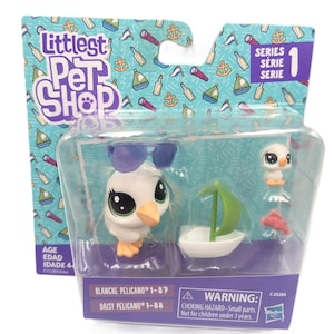 Puede incluir: Un set de juguetes Littlest Pet Shop Serie 1 que incluye una figura de pelícano blanco con gafas de sol moradas, un pequeño velero blanco y verde, una diminuta figura de pájaro blanco y un pequeño pez rojo, en su empaque original con texto.