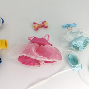 Puede incluir: Un conjunto de ropa para muñecas que incluye un vestido de lunares rosa y blanco, un atuendo azul y blanco, una diadema azul, un tutú azul y blanco, un lazo azul y dos pares de zapatos de ballet azules.