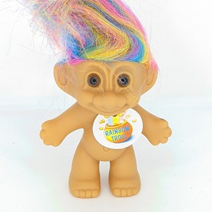 May include: A small, plastic rainbow troll doll with a colorful, fluffy hairpiece. The doll is wearing a white sticker with the words "Rainbow Troll" in a yellow circle.