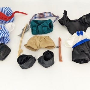 Puede incluir: Un conjunto de ropa y accesorios para muñecas, que incluye una falda a rayas azules y blancas, un sombrero a rayas rojas y blancas, un chaleco verde, pantalones cortos de color canela, un vestido negro, una camisa blanca con una corbata de moño azul, un violonchelo negro y botas negras.