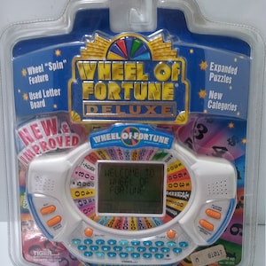 May include: A white handheld electronic game based on the Wheel of Fortune game show. The game features a spinning wheel with dollar amounts, a letter board, and a digital display. The game is packaged in a clear plastic clamshell with a blue and gold background. The packaging features the text "Wheel of Fortune Deluxe" and "New & Improved".