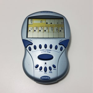 May include: A blue and silver handheld electronic game called "Card Stacks On-Deal-Draw" by Radica. The game has a small LCD screen with a card game display and buttons for "Deck", "Suits", "New Game", "Reset", "Sound", "Undo", "High Score", and "Timer".