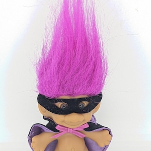 Puede incluir: Un muñeco troll morado con una máscara negra y una capa morada con un lazo rosa.