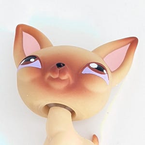 Könnte beinhalten: Ein hellbraunes Chihuahua-Spielzeug mit großen Ohren und einem lächelnden Gesichtsausdruck. Das Spielzeug hat eine hellrosa Nase und Innenohren mit lila Akzenten um die Augen. Das Spielzeug steht auf seinen Hinterbeinen.