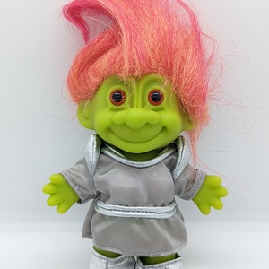 May include: A vintage Troll doll with bright pink and orange hair. The doll has a green face, large eyes, and a smiling expression. It is wearing a gray outfit with silver accents, including a matching skirt and boots.