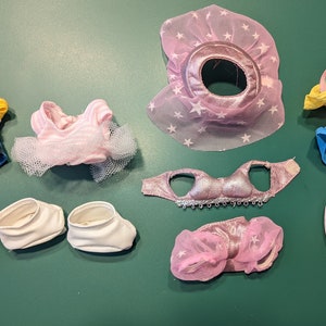 Puede incluir: Una colección de 10 prendas y accesorios para muñecas, que incluyen un tutú rosa, un atuendo deportivo azul y amarillo, un atuendo de bailarina rosa y blanco, un sombrero rosa y blanco en forma de estrella, un tutú rosa y blanco, un sujetador rosa y blanco, un par de zapatos de ballet blancos y un par de zapatillas blancas.
