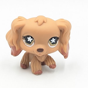 Könnte beinhalten: Ein hellbrauner Spielzeughund mit großen Augen, die weiße blütenförmige Akzente aufweisen. Der Hund hat Schlappohren und eine kleine braune Nase. Das Spielzeug ist ein Sammlerstück, wahrscheinlich für Kinder oder Sammler.