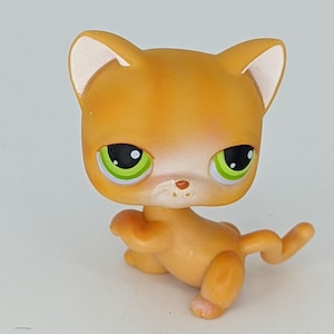 Puede incluir: Gato de juguete de plástico naranja y blanco con ojos verdes. El gato está sentado sobre sus patas traseras con las patas delanteras levantadas.