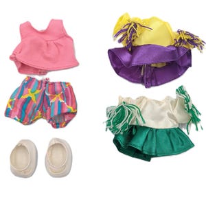 Puede incluir: Un conjunto de ropa de muñeca, que incluye una camiseta rosa, pantalones cortos estampados y zapatos blancos. Dos conjuntos adicionales presentan una camiseta amarilla con falda morada y una camiseta blanca con falda verde. Cada conjunto tiene accesorios a juego.